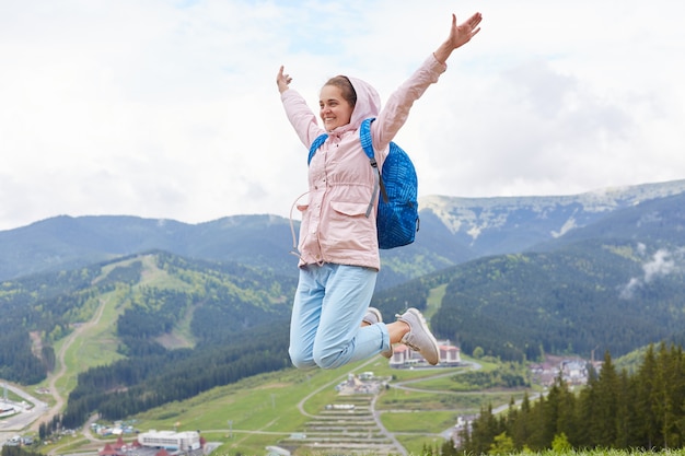 Tiro al aire libre de la mujer saltando alto, puerto de senderismo en vacaciones. Hembra que lleva la chaqueta rosada y la mochila azul aisladas sobre las montañas. Concepto de viaje, aventura y recreación activa.
