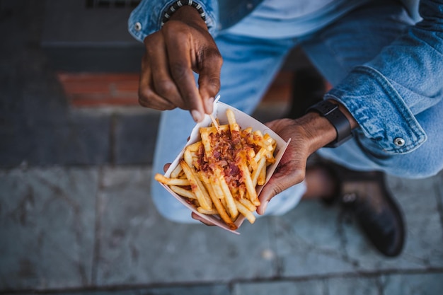 Tiro de ángulo alto de un hombre comiendo papas fritas con queso cheddar y tocino sentado en la calle