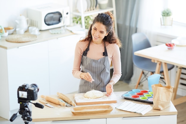 Tiro de ángulo alto horizontal de joven mujer caucásica con pelo rizado de pie en la mesa de la cocina preparando masa en la cámara