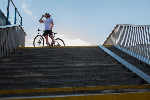 Tiro de ángulo bajo de un ciclista musculoso masculino bebiendo agua, de pie cerca de su bicicleta en el día asummer, espacio de copia