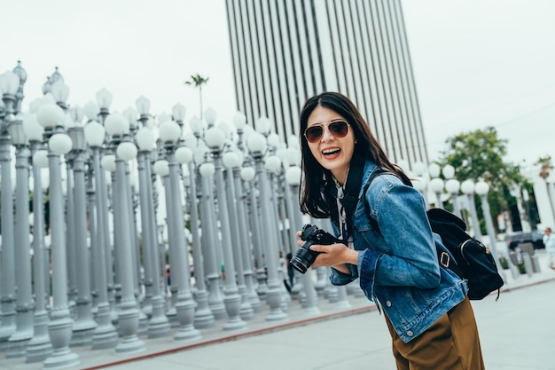 tiro de ángulo holandés mujer coreana que sostiene una cámara digital se ríe de buena gana frente a una instalación de arte en los Ángeles. una turista asiática con gafas de sol está mirando la lente de la cámara con una gran sonrisa.