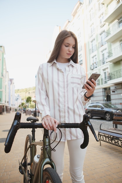 Tiro de ángulo bajo vertical de una mujer con teléfono inteligente, caminando en la ciudad con su bicicleta