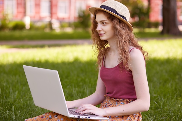 Tiro ao ar livre da bela jovem sentada no parque com o laptop nas pernas, passando o dia de verão trabalhando ao ar livre, olhando para a distância, vestindo camiseta, saia e chapéu, garota gosta de trabalho on-line ao ar livre.