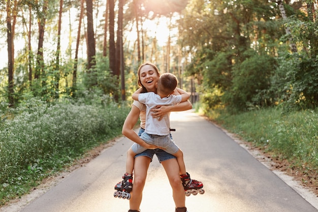 Foto tiro ao ar livre da mãe com o garotinho patinando no parque de verão, mãe segurando a criança nas mãos, abraçando e sorrindo com felicidade, aproveitando o tempo com o filho.