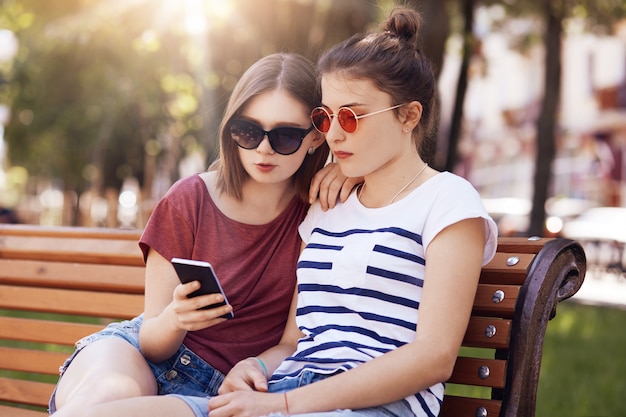 Tiro ao ar livre de duas belas fêmeas adolescentes olhar atentamente para telefone inteligente