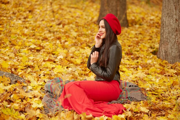 Tiro ao ar livre de jovem atraente garota bonita boina vermelha, calças e jaqueta de couro, sentado perto de árvores em amarelo leavesin