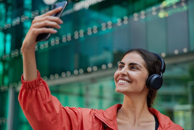 Tiro ao ar livre de mulher morena ouve música em fones de ouvido clica em fotos de selfie em sorrisos de smartphone amplamente usa jaqueta leva estilo de vida saudável usa tecnologias modernas. Pessoas e estilo de vida ativo