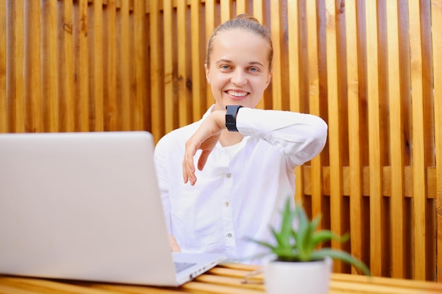 Tiro ao ar livre de mulher sorridente vestindo camisa branca e óculos sentado no escritório com paredes de madeira trabalhando no laptop e usando relógio inteligente expressando emoções positivas