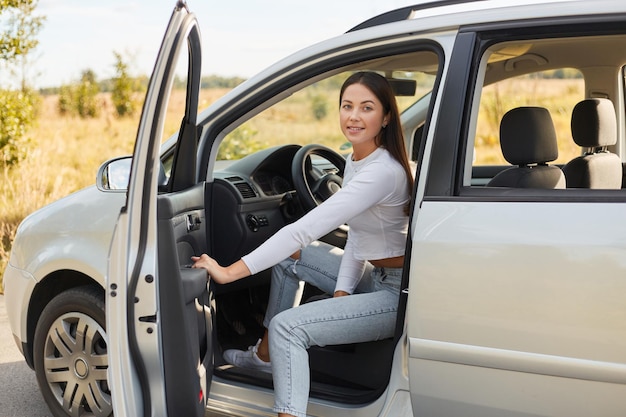 Tiro ao ar livre de sorridente mulher satisfeita com cabelo escuro saindo do carro com feliz expressão facial positiva feminina vestindo jeans e camisa branca