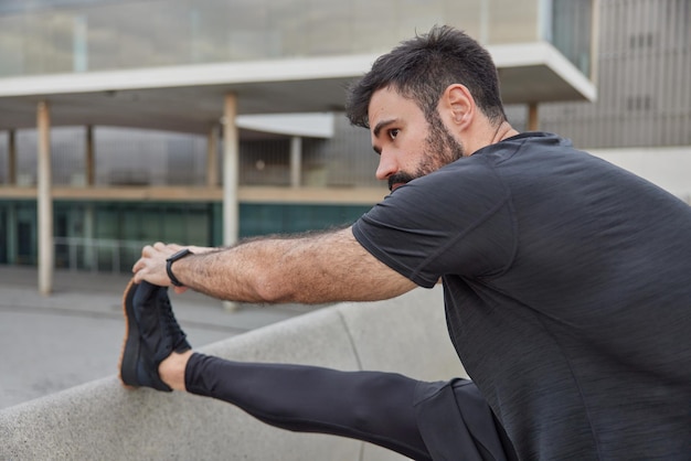 Tiro ao ar livre de um homem barbudo em roupas esportivas faz exercícios de alongamento antes do treinamento ficar pronto para a rotina de corrida focada na distância com expressão motivada. pessoas e conceito de treino