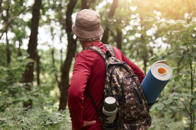 Tiro ao ar livre do velho homem tendo saco com garrafa térmica e almofada para dormir, usando chapéu bege e moletom vermelho, procurando aventuras na floresta sozinha, gosta de viajar e fazer caminhadas.