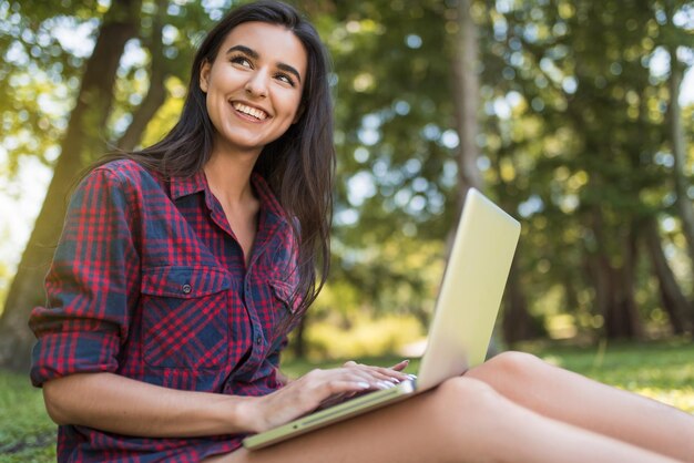 Tiro atraente de jovem mulher caucasiana feliz vestindo camisa xadrez, sentado na grama na frente do laptop pc trabalhando em suas compras de computador portátil e comunicação sem fio