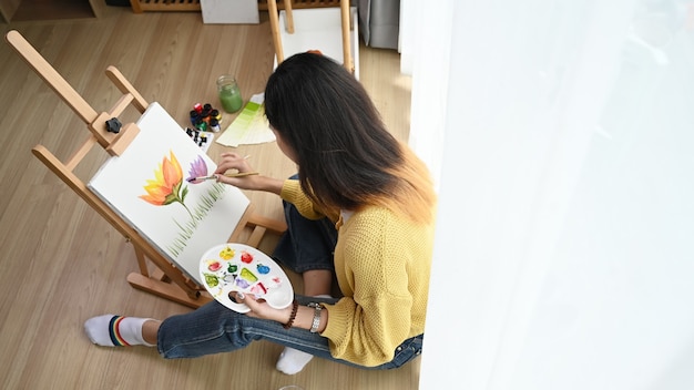 Tiro a la cabeza de una joven artista en suéter amarillo con paleta en la mano está pintando sobre lienzo en el estudio.