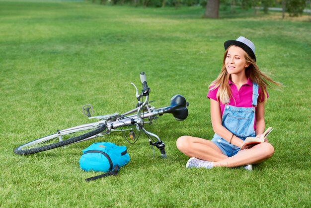 Tiro de un ciclista femenino joven hermoso que descansa sobre la hierba en el parque después de completar un ciclo que lee un libro que sonríe felizmente recreación afición literatura inteligencia felicidad estilo de vida concepto de verano.