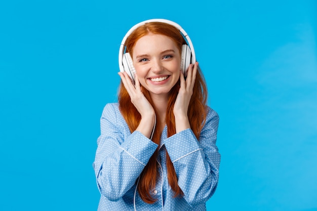 Tiro con la cintura alegre alegre bastante astuta niña caucásica escuchando música en auriculares grandes, sonriendo complacido, mirando amigos solteros, vistiendo ropa de dormir, parado pared azul encantado