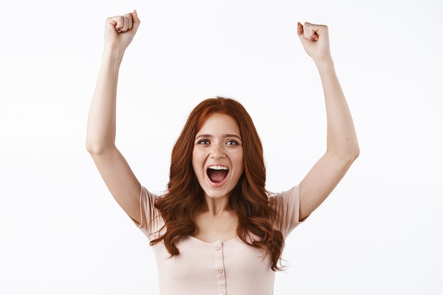 Tiro de cintura para arriba alegre y entusiasta chica pelirroja bombea su puño con alegría, celebrando noticias increíbles, sonriendo complacida, gritando sí celebrando, ganando, convirtiéndose en campeona en el juego, apoyando al equipo