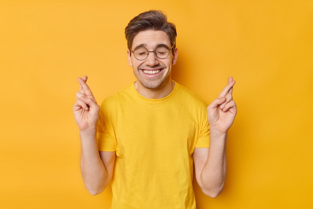 Tiro de cintura para arriba de cabello oscuro positivo hace que el deseo preciado ruega por suerte espera que sucedan buenos pensamientos usa gafas redondas y una camiseta casual aislada en fondo amarillo Deseo deseo superstición