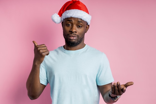 Tiro de cintura para arriba de un joven afroamericano sin afeitar satisfecho con sombrero de Santa, hablando con alguien, pidiendo ver algo interesante, apuntando con el pulgar a un lado, fondo rosa aislado.