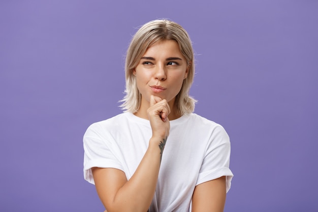 Tiro de cintura para arriba de mujer ambiciosa joven creativa inteligente con corte de pelo rubio y piel bronceada entrecerrando los ojos y sonriendo con satisfacción mirando a la izquierda mientras sostiene la mano en la barbilla pensando en plan o decisión