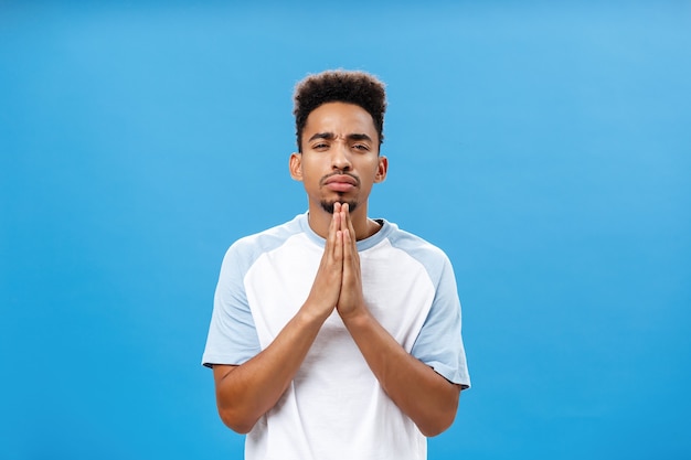 Tiro de cintura para arriba de novio afroamericano guapo intenso preocupado con barba tomados de la mano en oración frunciendo el ceño mirando pensativo a la cámara haciendo cálculos importantes sobre la pared azul. Copia espacio