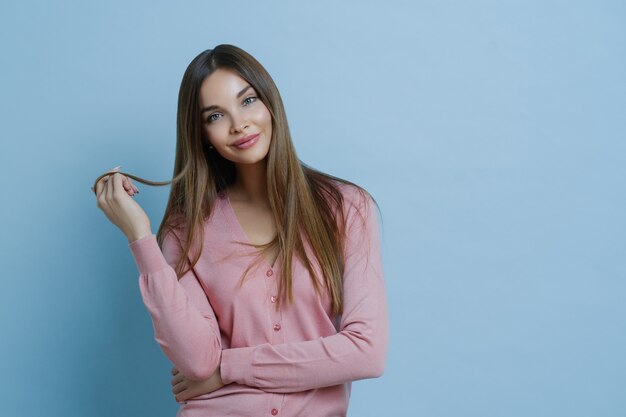 El tiro de la cintura de una mujer joven de aspecto agradable tiene un hermoso cabello largo, inclina la cabeza, se viste con un jersey informal, tiene una piel limpia perfecta