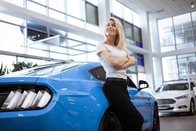 Tiro de ângulo baixo do motorista do sexo feminino deslumbrante posando com seu novo automóvel no salão da concessionária.