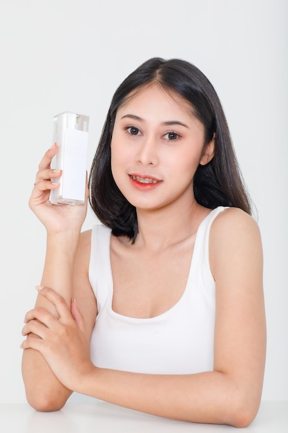 Tiro de beleza retrato do modelo feminino milenar jovem asiático de cabelo preto curto em camiseta regata, segurando o pacote de garrafa de creme facial de soro natural na mão sobre fundo branco.