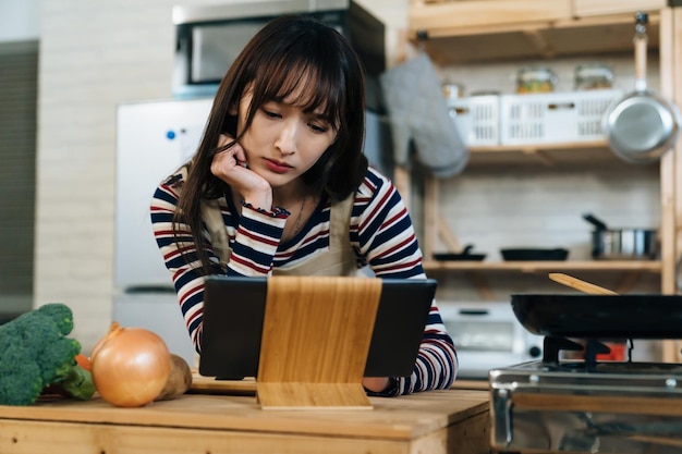 tiro de closeup de uma noviça de cozinha feminina asiática confusa apoiando seu rosto e inclinando-se perto da almofada elétrica enquanto lê receitas on-line para aprender a cozinhar em casa.