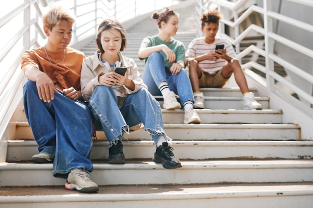 Tiro de comprimento total arejado de diversos adolescentes usando smartphones ao ar livre enquanto está sentado em escadas de metal