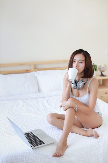 Tiro de comprimento total de linda mulher asiática adulta vestido branco, sentado na cama.