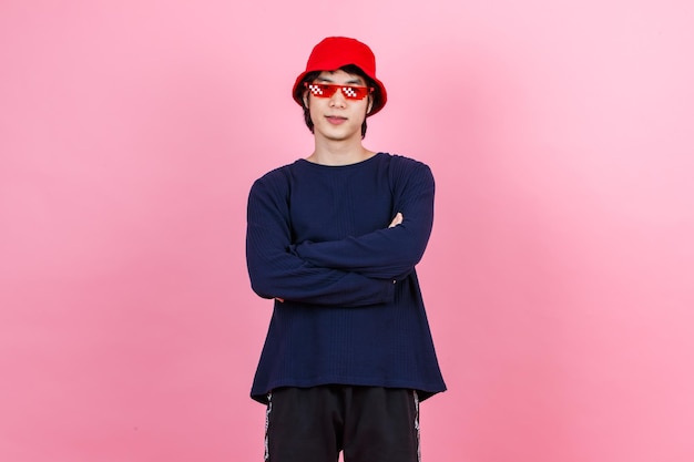 Tiro de estúdio de retrato asiático jovem urbano moda adolescente modelo masculino em roupa de estilo de rua camisa de manga longa chapéu de balde vermelho óculos engraçados em pé braços cruzados sorrindo olhe para a câmera no fundo rosa