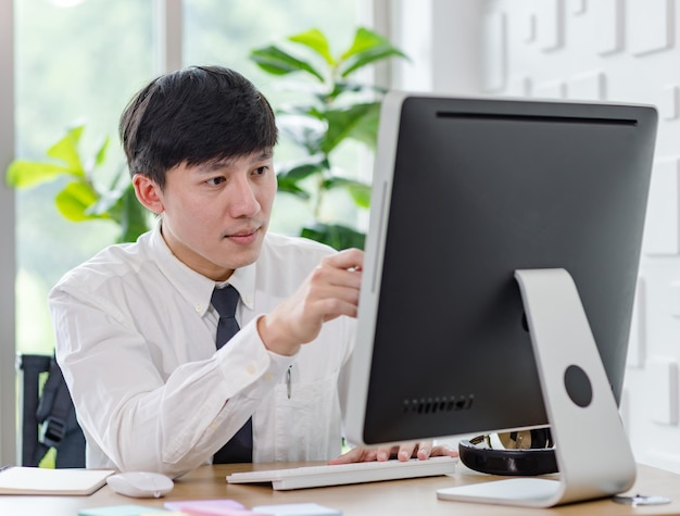 Tiro de estúdio de retrato de empregado de empresário masculino bem sucedido profissional asiático na camisa formal com gravata sentado olhar para a câmera na mesa de trabalho com mouse de teclado de monitor de computador e artigos de papelaria.