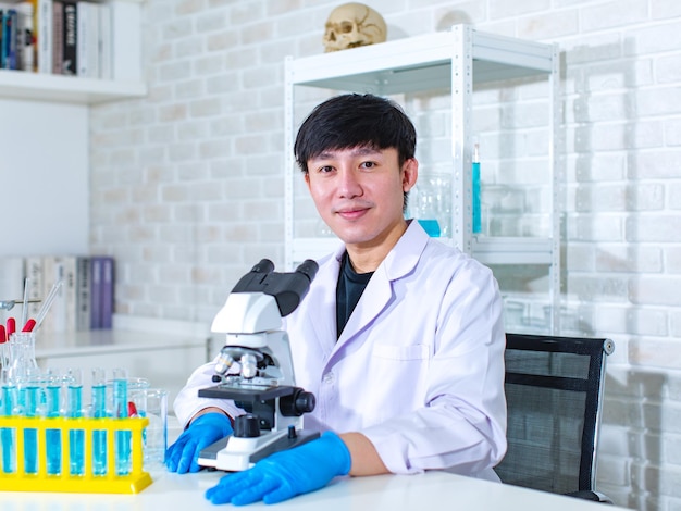 Tiro de estúdio de retrato de jovem cientista masculino profissional asiático no jaleco branco sentado sorrindo olhar para a câmera atrás da mesa de trabalho do laboratório cheia de amostras de frascos de tubos de ensaio e microscópio.