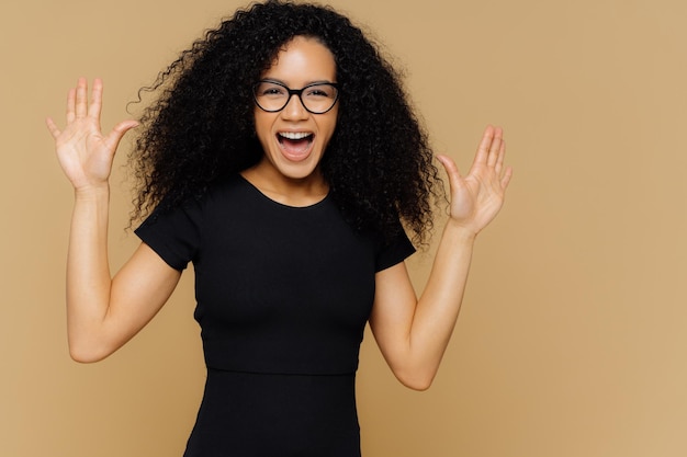 Tiro de metade do comprimento da mulher feliz e satisfeita dança e se move positivamente, levanta as palmas das mãos, exclama com felicidade, usa camiseta preta, óculos, isolado na parede bege. Modelos femininos animados se movem