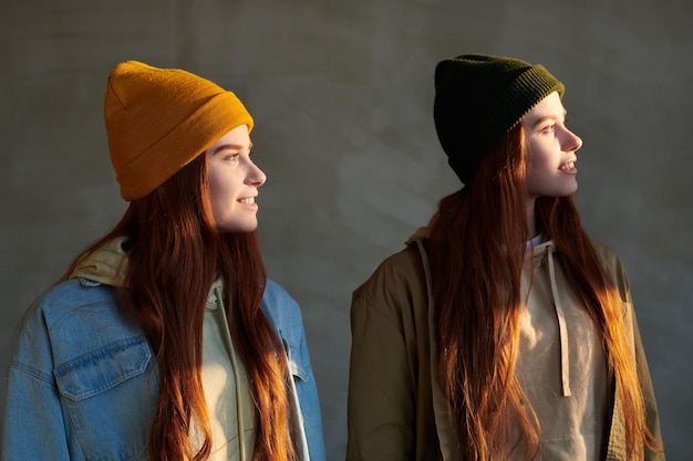 Tiro de retrato horizontal médio de duas mulheres jovens com longos cabelos vermelhos vestindo roupas casuais e bonés olhando para longe sorrindo