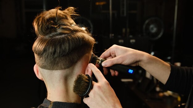 Tiro de um barbeiro bonito dando um corte de cabelo para seu cliente usando aparador Serviço de cabeleireiro em uma barbearia moderna em um raio escuro com luz quente vista traseira