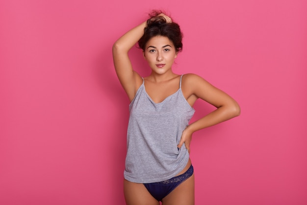 Tiro do modelo de roupa interior feminina. jovens se encaixam mulher vestindo camiseta de algodão cinza e calcinha azul, fêmea atraente fica contra a parede rosa, mantendo hnads em seus cabelos escuros, garota com corpo perfeito.