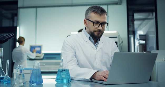 Tiro do retrato do homem caucasiano de óculos e roupão branco, trabalhando no computador portátil no laboratório ao lado com um microscópio.