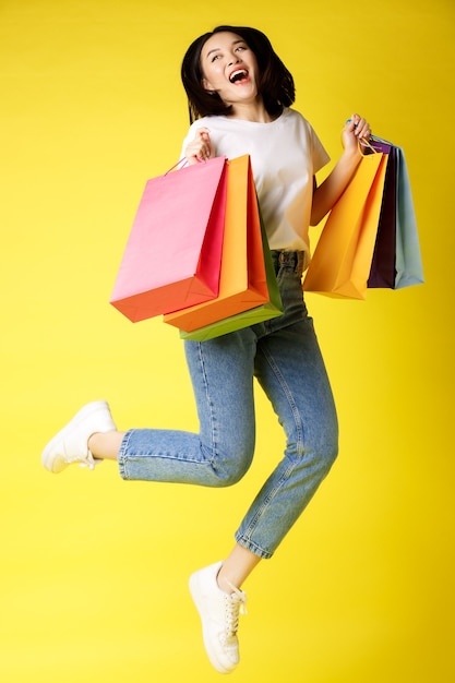 Tiro em tamanho real de linda mulher asiática pulando com sacolas de compras e sorrindo feliz, fundo amarelo.