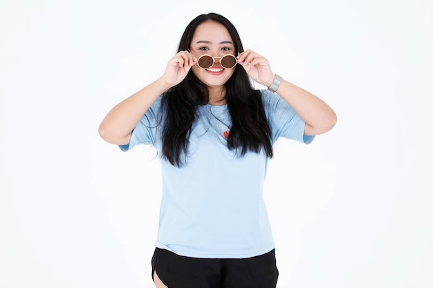 Tiro de estudio de retrato de modelo femenino de pelo largo negro sano regordete asiático rechoncho en camisa casual azul permanente mirada sonriente a cámara sosteniendo tocar quitar gafas de sol vintage sobre fondo blanco.
