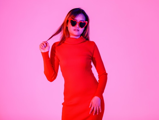 Tiro de estudio de retrato de modelo de moda femenina confiada asiática en vestido de cuello alto de manga larga rojo de moda casual con gafas de sol de lente negra en forma de corazón de pie sexy posando sobre fondo blanco.