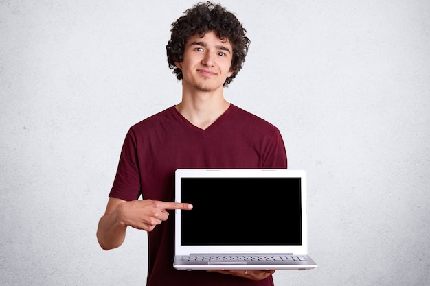 Tiro del hombre hermoso sonriente que lleva la camiseta marrón casual que coloca y que señala el dedo en la pantalla en blanco del ordenador portátil con la expresión facial plasant.