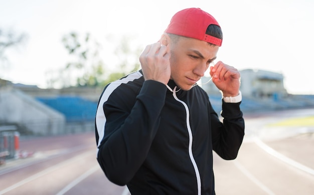 Tiro horizontal de un apuesto joven atlético usando auriculares mirando hacia abajo en el estadio Atractivo corredor escuchando música durante el trote matutino y corriendo al aire libre Gente deportiva y estilo de vida