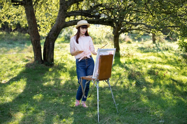 Tiro horizontal de cuerpo entero de una mujer caucásica bastante joven, con sombrero de paja, jeans y camisa rosa, pintura sobre lienzo afuera en el parque.