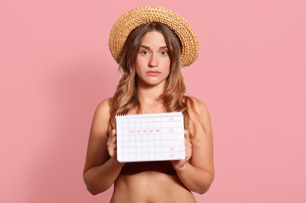 Tiro horizontal da bela jovem europeu feminino vestido com chapéu de palha e maiô, segurando o calendário do período, tem expressão infeliz, tem a menstruação.