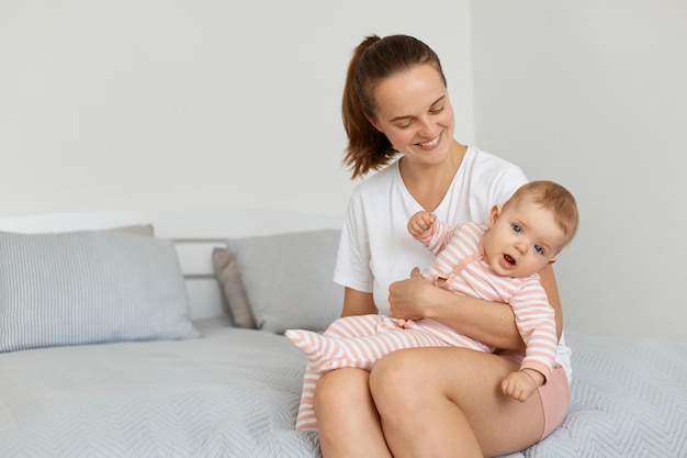 Tiro horizontal da jovem mãe adulta feliz vestindo camiseta casual branca, sentado na cama no quarto com a mamãe bebê da criança olhando para a criança com um sorriso encantador, feliz por passar tempo com seu filho