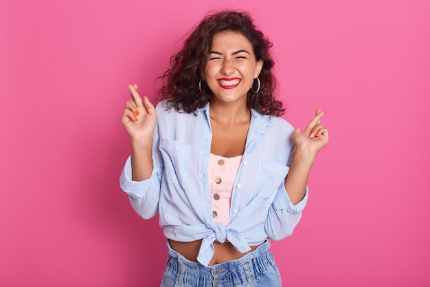 Tiro horizontal da jovem mulher atraente mantém os dedos cruzados e os olhos fechados