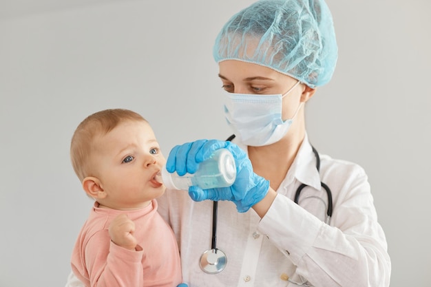 Tiro horizontal da médica com estetoscópio sobre o pescoço usando um vestido, boné médico, máscara cirúrgica, em pé com a menina nas mãos e alimentando a criança de garrafa.