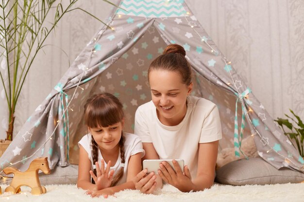 Tiro horizontal da mulher deitada no chão perto da família wigwam infantil usando telefone celular para fazer uma videochamada ou transmitir ao vivo para o blog da família criança acenando com a mão passando um tempo juntos