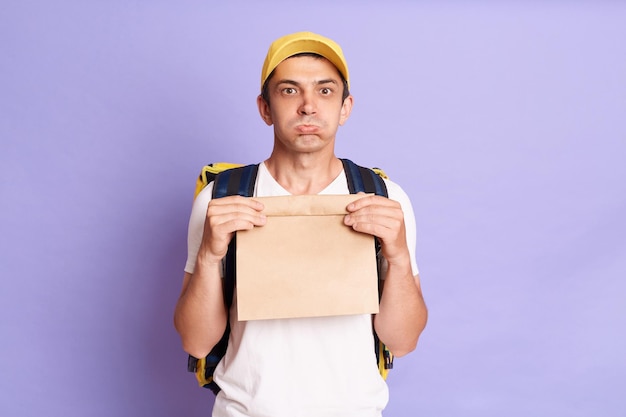 Tiro horizontal de entregador triste chateado segurando o pacote de papel com comida isolada no fundo violeta expressando tristeza olhando para a câmera com lábios de beicinho